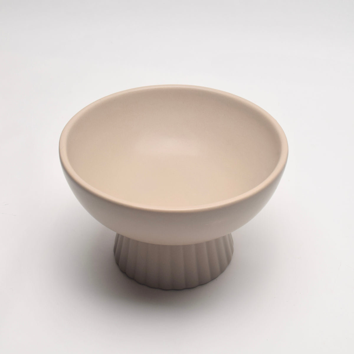 A minimalist ceramic pedestal bowl in a warm cream-beige color, featuring a fluted base and a smooth, rounded interior.