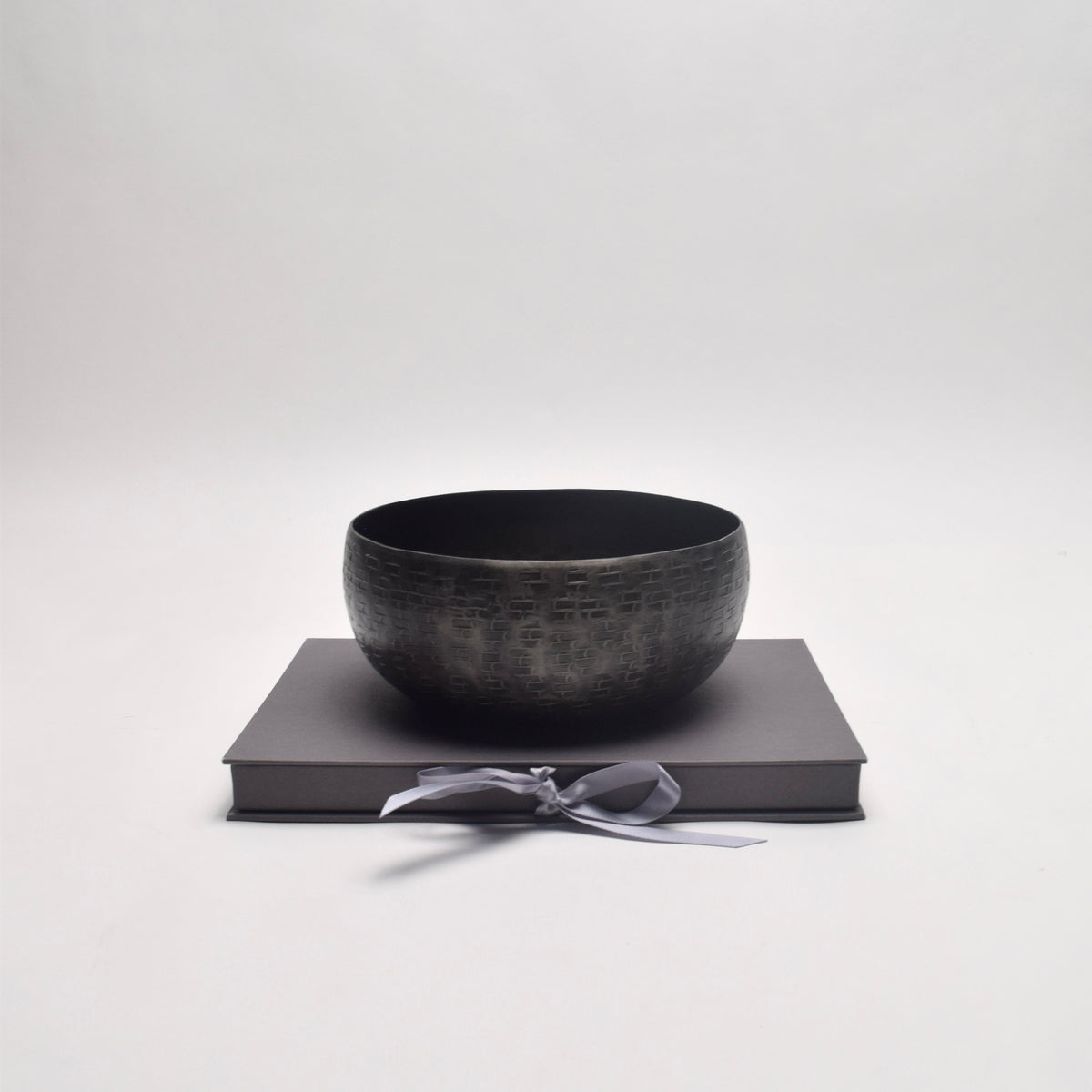 A dark textured metal bowl displayed on a dark grey rectangular box tied with a grey ribbon for shelf-styling