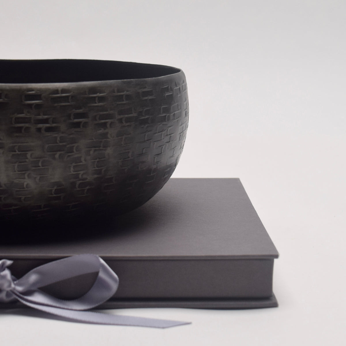 A dark textured metal bowl resting on a grey presentation box tied with a light gray ribbon, set against a clean light gray background.