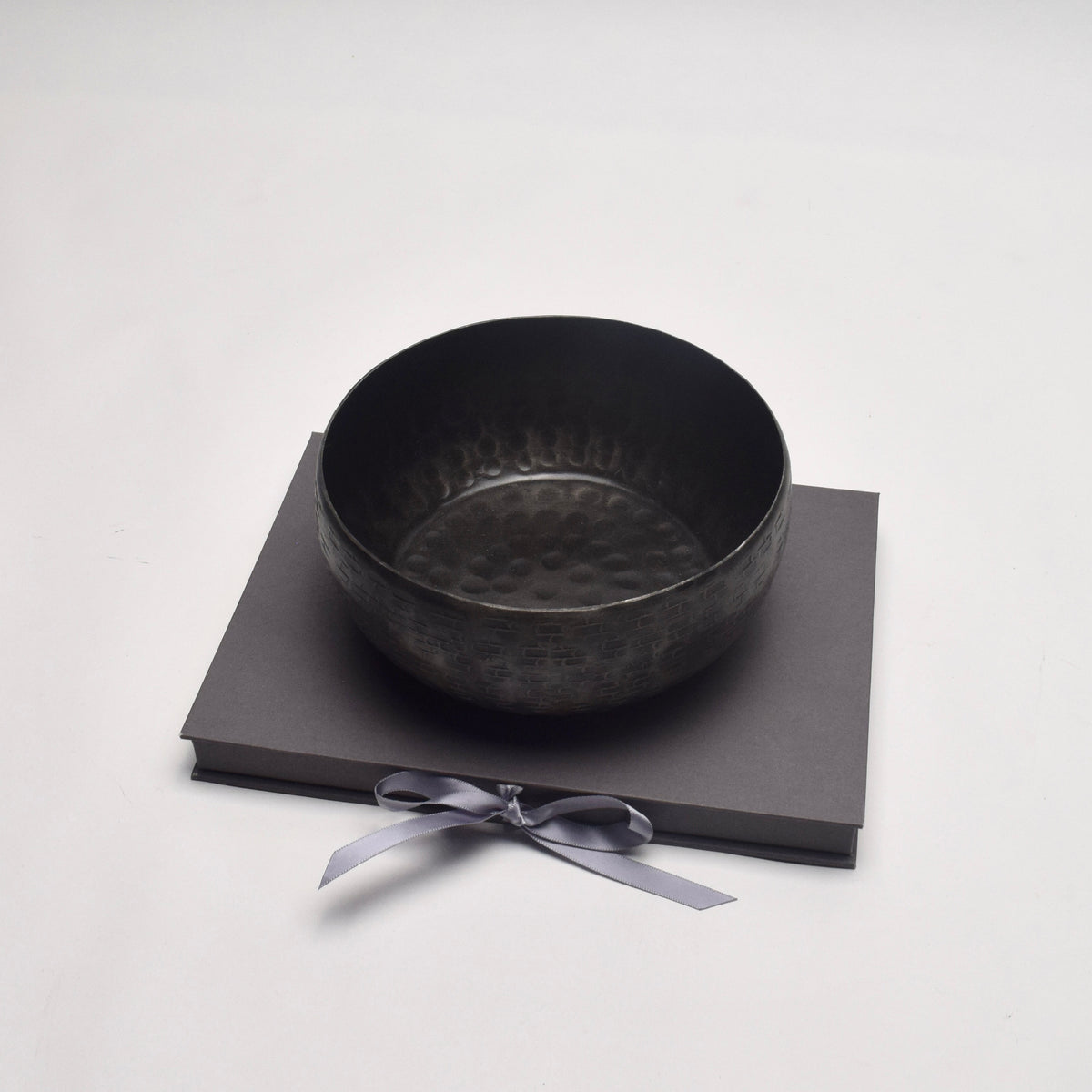 A dark hammered metal bowl displayed on top of a dark grey presentation box tied with a light grey ribbon, set against a plain light grey background.