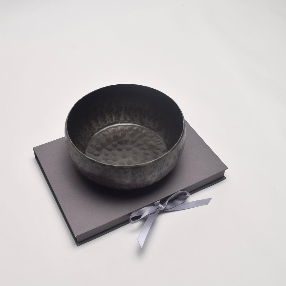 A dark grey hammered metal bowl resting on a square dark grey box tied with a light gray ribbon, photographed at above angle
