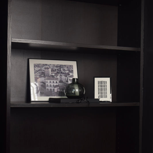 A dark wood shelving unit with two middle shelves displaying a black and white framed cityscape photograph, a small dark glass vase, and a white rectangular art print.