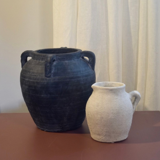 Two ceramic vessels, a large dark charcoal-black four-handled vase and a smaller textured off-white jug, displayed on a reddish-brown surface against a cream curtain backdrop.