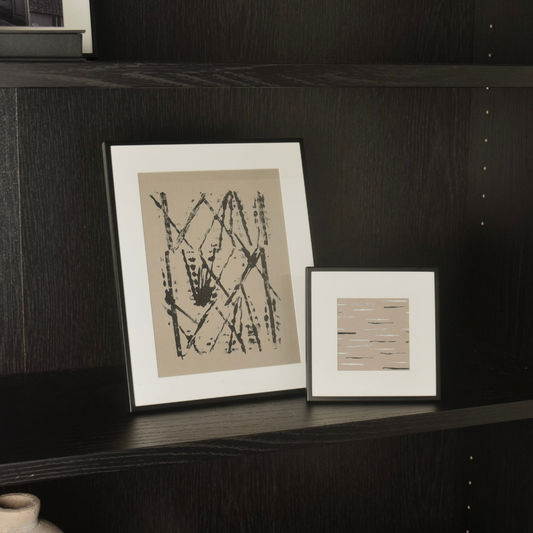 Two framed abstract art prints displayed on a dark wood shelf. The larger print features black brushstrokes on a beige background and the smaller print has horizontal lines in beige and brown tones.