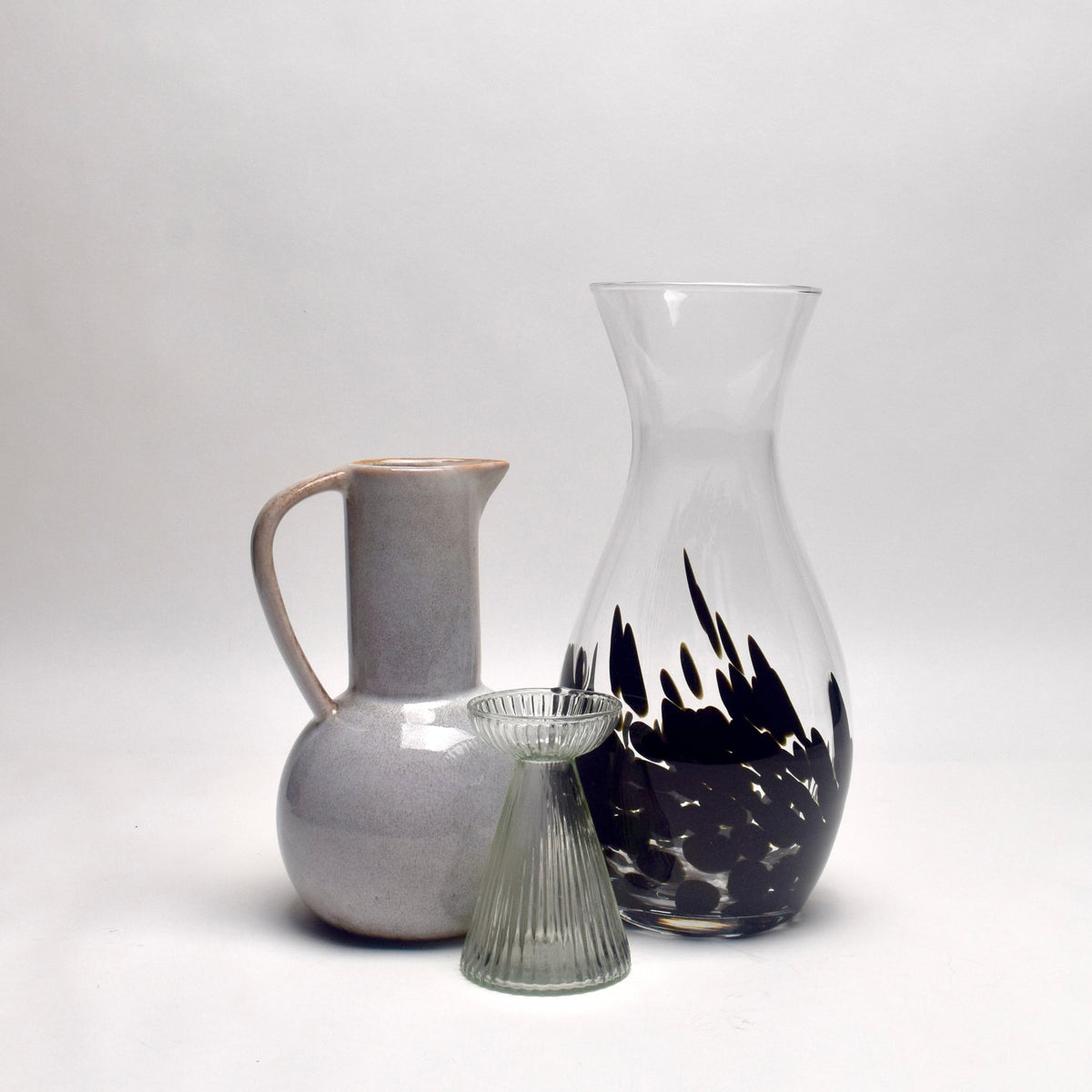 Three glass and ceramic vessels on a seamless light gray studio background: a matte light gray pitcher with a handle, a clear glass carafe with a black abstract flame-like pattern near the base, and a small ribbed clear glass vase in front.