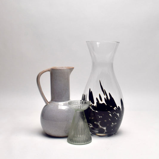 Three glass and ceramic vessels on a seamless light gray studio background: a matte light gray pitcher with a handle, a clear glass carafe with a black abstract flame-like pattern near the base, and a small ribbed clear glass vase in front.