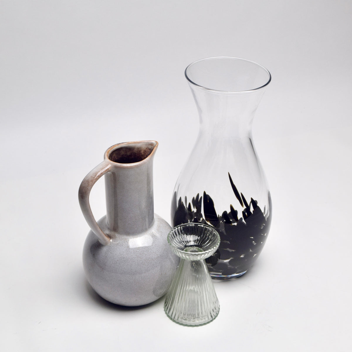 Three glass and ceramic vases on a seamless light gray studio background: a tall clear glass carafe with a black leaf-like pattern, a pale gray glazed pitcher, and a small ribbed clear glass vase.