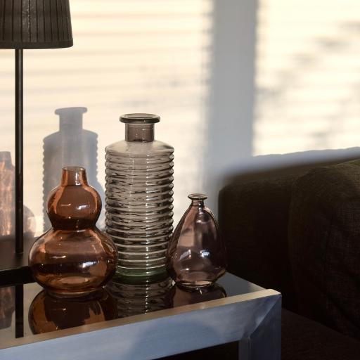 Three decorative glass vases in amber, smoky grey, and translucent brown tones arranged on a reflective side table in a softly lit room with window blinds and a dark sofa in the background.