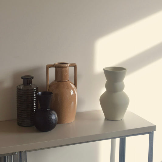 A set of four minimalist ceramic vases in black, orangey-brown, and cream, arranged on a  console table against a warm beige wall with soft diagonal sunlight.